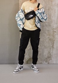 Giacca a vento con motivo astratto blu e bianco, t-shirt beige con logo, pantaloni cargo neri con tasche con zip e sneakers grigie.