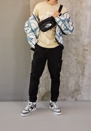 Een pufferjack met een blauw-wit abstract patroon, een beige t-shirt met logo, zwarte cargobroek met ritszakken en grijze sneakers.