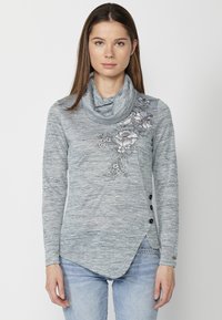 Top à col châle à manches longues en tissu tricoté gris clair, avec une broderie florale sur une épaule et trois boutons noirs sur l'ourlet latéral.