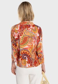 Blouse à imprimé cachemire multicolore avec manches trois-quarts, présentant des rouges, oranges et jaunes vibrants ainsi que des motifs complexes sur fond blanc.