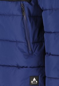 Marinblå puffy-jacka med svarta accenter, med en zip-försedd bröstficka och en logotyp-patch på den nedre framsidan. Mönstrad quiltad design.