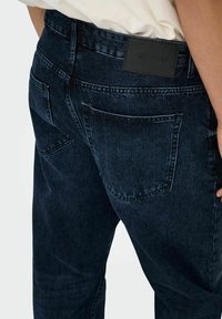 Jeans de mezclilla azul oscuro con un corte ajustado, que presentan una etiqueta de cuero en la cintura y dos bolsillos traseros con detalles de costura visibles.