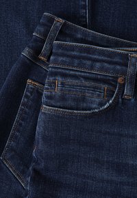 Jeans de mezclilla azul oscuro con costuras naranjas, diseño de cinco bolsillos y una textura suave. Detalles notables incluyen un pequeño bolsillo para monedas.