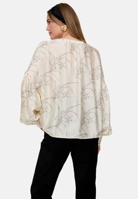 Cremefarbene Bluse mit floralem Linienzeichnungsmuster, weiten Ärmeln und lockerem Schnitt. Kombiniert mit schwarzen Hosen für einen Kontrast.