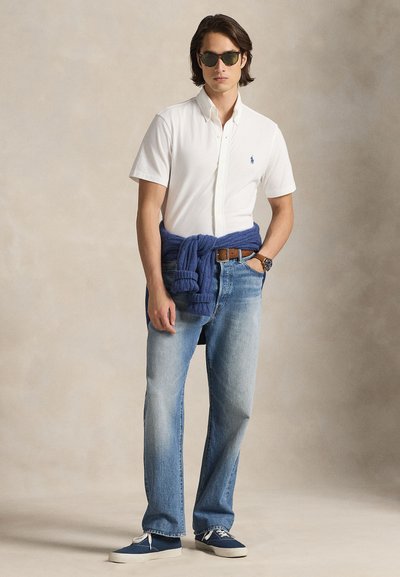 Polo branca de manga curta, jeans azul claro, suéter azul amarrado na cintura, sapatos azuis e óculos de sol. Estilo casual clássico sem padrões.