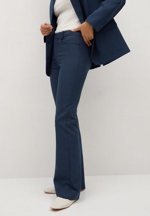 Žena v tmavě modrých upravených kalhotách a sladěném saku přes bílý top, upravující sako, s jednou rukou v kapse, stojící na bílém podlaze.