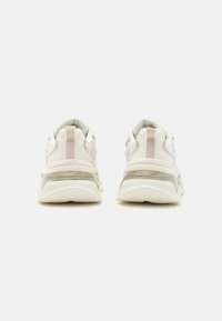 Jasne buty sportowe z gładką materiałową cholewką, przezroczystą podeszwą i teksturowanymi detalami na pięcie i bokach.