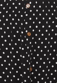 Mustaa kangasta valkoisilla pilkuilla, jossa on teksturoitu pinta. Edessä on ruskeita pyöreitä nappeja kiinnitystä varten.