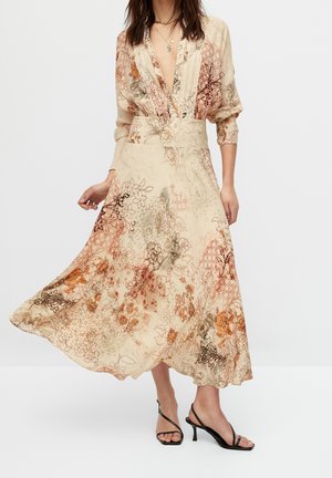 Woman wearing a flowing, long floral dress with rolled sleeves and black strappy heeled sandals, standing on a plain background.