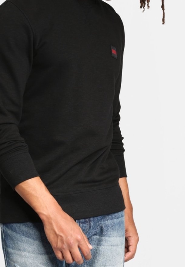 Sweatshirt noir à manches longues avec un col rond, tissu texturé, et un petit écusson logo rouge et noir sur la poitrine gauche. Porté avec un jean bleu.