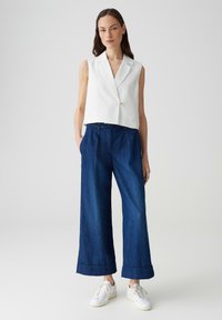 Blazer bianco senza maniche con un solo bottone, abbinato a jeans larghi blu scuro. Accentuato da risvolti e sneakers bianche.