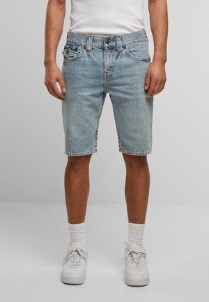 Shorts in jeans leggeri di colore azzurro con vita media, design a cinque tasche, cuciture visibili e una texture leggermente usurata. Indossati con una camicia bianca e sneaker.