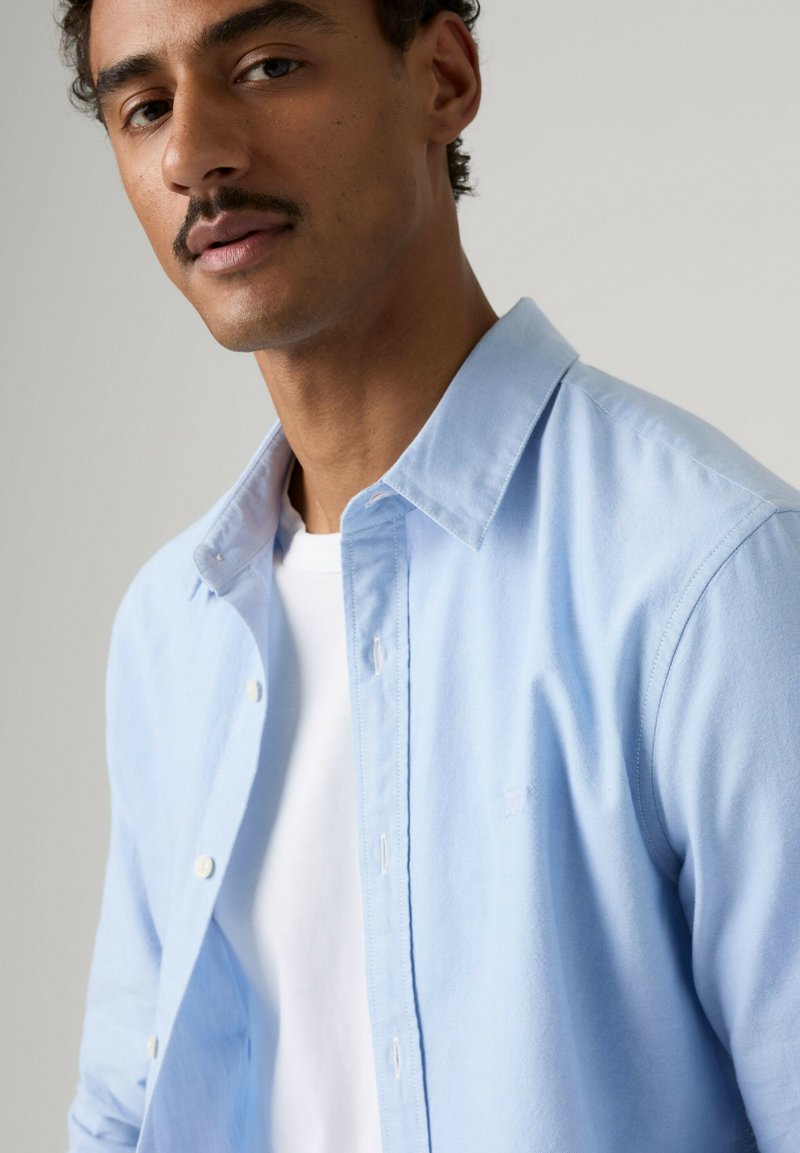 Mand med kort krøllet hår og mustache iført en lyseblå skjorte over en hvid T-shirt mod en ensfarvet baggrund.
