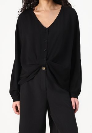 Vrouw met een zwarte blouse met lange mouwen, V-hals en knopen, gestrikt in de taille, gecombineerd met zwarte broek met hoge taille en zichtbare knopen.