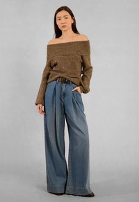 Maglione a spalle scoperte marrone in maglia con riflessi metallici, abbinato a jeans in denim blu a vita alta e gamba larga. Accentato da una cintura nera.