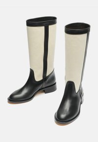 Botas hasta la rodilla con punta y tacón de cuero negro, con un caña de lienzo crema y un acento de cuero negro en el centro.
