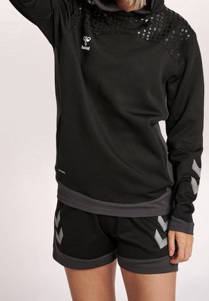 Person iført sort sportstøj med mønstrede skuldre og chevron-detaljer på ærmer og shorts, stående med den ene arm hævet.