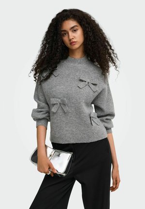 Femme aux cheveux bouclés portant un pull gris orné de nœuds et un pantalon noir, tenant une pochette argentée dans sa main droite.