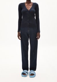 Osoba oblečená v tmavě modrém přízí žebrovaném kardiganu na knoflíky, volných tmavě modrých džínách a světle modrých chlupatých pantoflích s otevřenou špičkou, stojící před bílým pozadím.