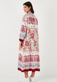 Vestido maxi floral con patrones vibrantes en rojo, rosa y verde sobre un fondo blanco, con mangas largas y un dobladillo burdeos.