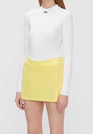 Kobieta ubrana w biały top z długim rękawem i podwyższonym kołnierzem z małym czarnym logo oraz żółtą, wysoką spódnicę mini z subtelnymi bocznymi zakładkami.