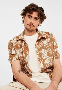 Brązowa koszula z krótkim rękawem, w białe kwiatowe wzory, z guzikiem z przodu, noszona na kremowym t-shircie. Model z widocznymi tatuażami, siedzący.
