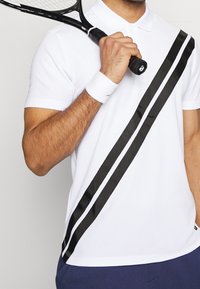 Bílá polo košile s černými saténovými pruhy šikmo přes přední stranu. Obsahuje klasický límec a krátké rukávy. Drží tenisovou raketu.