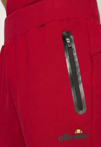 Rote Trainingshose mit einer schwarzen Reißverschlusstasche an der Seite, aus strukturiertem Stoff gefertigt, mit einem kleinen Logo nahe dem Saum.