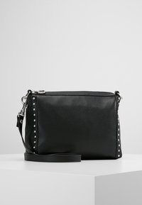 Sac bandoulière en cuir noir texturé avec des clous argentés le long des côtés et une bandoulière amovible, posé sur une surface blanche.