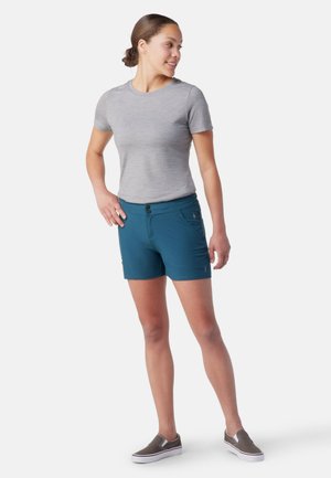 Türkisblaue Shorts mit glatter Textur, mit zwei Knöpfen und seitlichen Taschen. Kombiniert mit einem hellgrauen, figurbetonten T-Shirt und lässigen Schuhen.