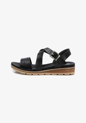Sandal de cuero negro con correas perforadas, hebilla ajustable y una suela apilada y texturizada en tonos marrón y claro. Presenta un diseño plano.