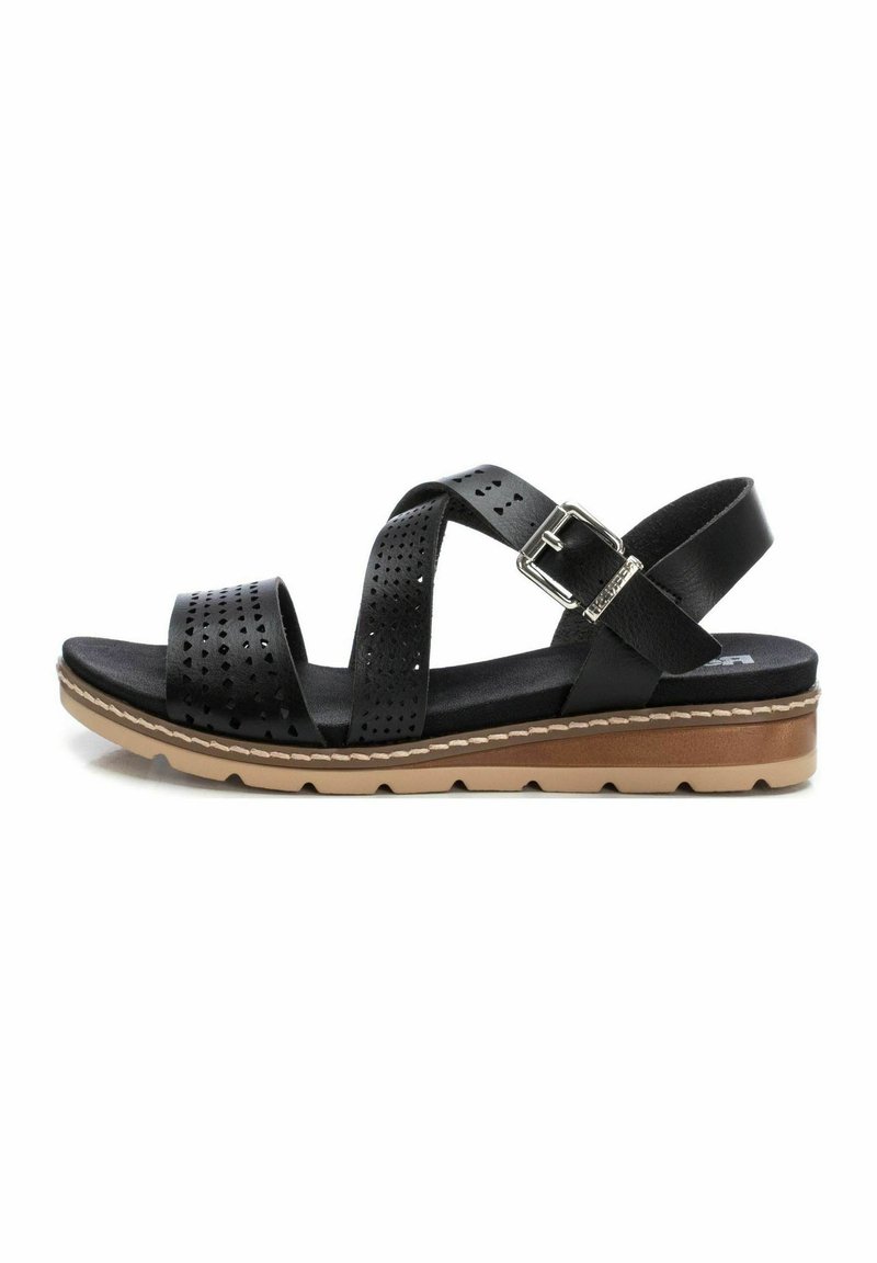Sandal de cuero negro con correas perforadas, hebilla ajustable y una suela apilada y texturizada en tonos marrón y claro. Presenta un diseño plano.