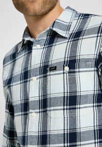 Man i blå och vit rutigt skjorta med knappar, bröstficka och liten svart "Lee" etikett.