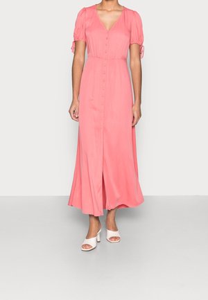 Woman wearing a coral pink buttoned midi dress with puff sleeves and white open-toe heeled sandals, standing on a plain background.