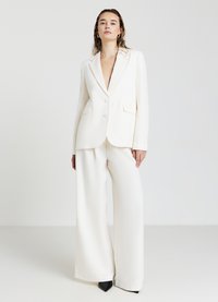 Blazer blanc ajusté avec un décolleté profond et deux poches avant, associé à un pantalon blanc à jambes larges et des escarpins noirs à bout pointu.