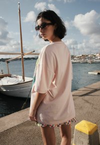 Prevelika roza majica s grafičnim vzorcem, obrobljena z večbarvnim karo zaključkom na spodnjem delu, nošena s temnimi sončnimi očali ob pristanišču.