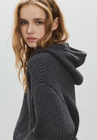 Szürke kötött kapucnis pulóver texturált mintával és csatlakoztatott kapucnival. Az anyag puhanak és vastagnak látszik, laza fazonban.