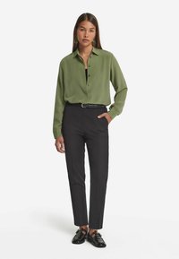 Olivgrüne Bluse mit langen Ärmeln und Knöpfen, kombiniert mit schwarzen engen Hosen und schwarzen Slippern. Schlichtes Design mit einem Gürtelakzent.