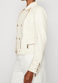 Veste en cuir crème à double boutonnage, fermeture éclair, quincaillerie dorée et détails de sangle aux poignets. Texture lisse, coupe ajustée.