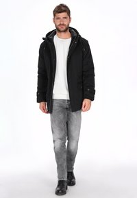 Chaqueta negra con capucha, cremalleras y botones, suéter gris debajo, jeans gris claro y botines negros. Textura de tela suave.