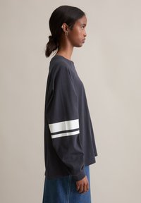 Donkergrijze langemouwenshirt met een ronde hals en witte strepen op de mouwen. Zachte stof en relaxte pasvorm. Gemaakt met een denim rok.