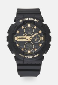 Orologio G-Shock nero con cinturino in gomma testurizzata, dettagli dorati, display digitale e lancette analogiche. Tra le caratteristiche più importanti è presente un'etichetta di protezione.