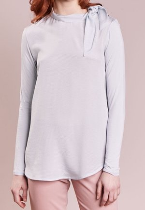 Mujer con una camiseta de manga larga gris claro con un detalle de nudo atado en el hombro izquierdo y pantalones rosa pálido sobre un fondo liso.