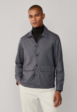 Chaqueta gris de botones con cuello, dos bolsillos frontales, corte entallado y textura suave, usada sobre un jersey de cuello alto negro.