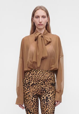 Femme portant un chemisier marron transparent avec un grand nœud et un pantalon taille haute à imprimé léopard, debout devant un fond uni.