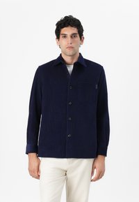 Camisa de pana azul marino con cuello puntiagudo, bolsillo en el frente y cierre de botones. Mangas largas con detalle en los puños. Tejido texturizado y ajuste casual.