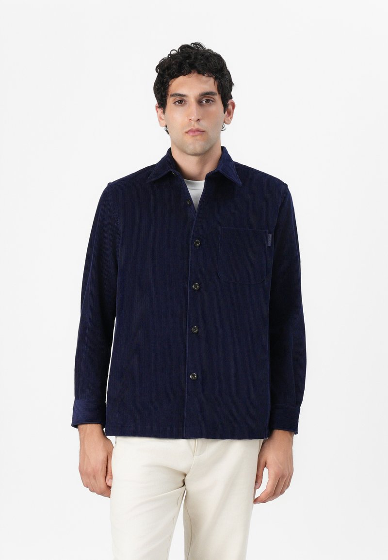 Camisa de pana azul marino con cuello puntiagudo, bolsillo en el frente y cierre de botones. Mangas largas con detalle en los puños. Tejido texturizado y ajuste casual.