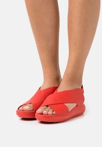 Sandali a zeppa rossi in pelle con cinturini incrociati, design con punta aperta e suola testurizzata. Presentano una base imbottita per maggiore comfort.