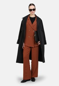 Un costume marron en deux pièces avec une veste croisée et un pantalon à jambes larges, associé à un long manteau noir et une pochette noire. Des lunettes de soleil foncées sont portées.