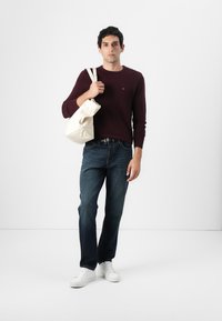 Pull à manches longues bordeaux, jean en denim bleu, baskets blanches, tenant un tote bag couleur crème. Fond uni, éclairage neutre.